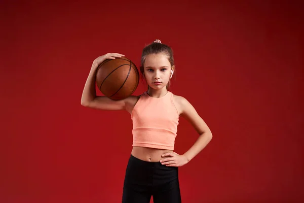 Oyun zamanı. Tatlı bir çocuk, kız sporla meşgul, basketbol oynarken kameraya bakıyor. Kırmızı arka planda izole edilmiş. Fitness, eğitim, aktif yaşam tarzı konsepti. Yatay çekim — Stok fotoğraf