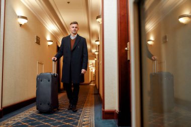Business travel. Middle-aged businessman in coat with suitcase walking along the hall, while looking for his room in a luxury hotel.