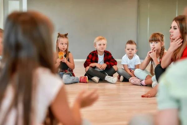 Communicating with children. Full length portrait of cute little boys and girls sitting on the floor gathered around their female dance teacher and listening her carefully. Dance school — 스톡 사진