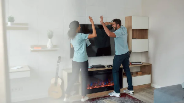 Good job. Two happy professional cleaners in uniform giving high five while working together in the living room. Young caucasian man and afro american woman wiping TV screen — Stockfoto