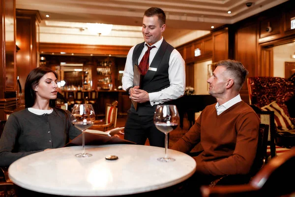 O serviço é o nosso principal objetivo. Garçom confiante dando cardápio aos clientes. Casal jantando no restaurante do hotel — Fotografia de Stock