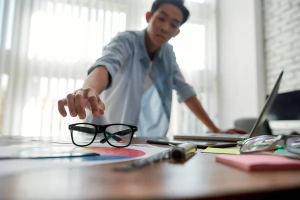 Taking care of my eyes. Busy young asian man in casual wear taking his eyeglasses before starting to work with laptop. Young designer working in the creative working space — Stock Photo, Image