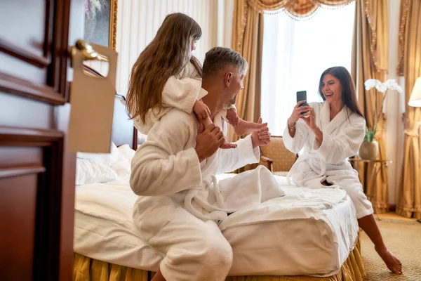 Sonríe. Padres e hija con batas blancas pasando la mañana juntos. Papá y el niño están posando para una foto sentados en la cama en una lujosa habitación de hotel. Familia, viajes, resort, concepto de vacaciones — Foto de Stock