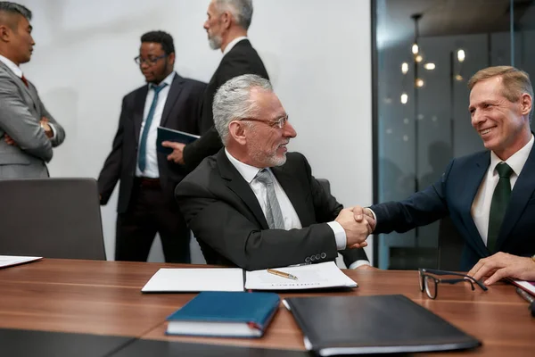 Dos hombres de negocios maduros felices en el desgaste clásico estrechando las manos después de la firma del contrato, mientras que tener una reunión multiétnica en la oficina moderna — Foto de Stock