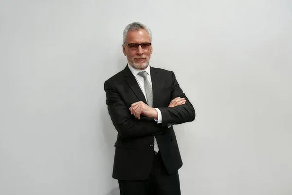 Looking perfect. Portrait of confident mature businessman in eyeglasses keeping arms crossed and looking at camera while standing against grey background — Stockfoto