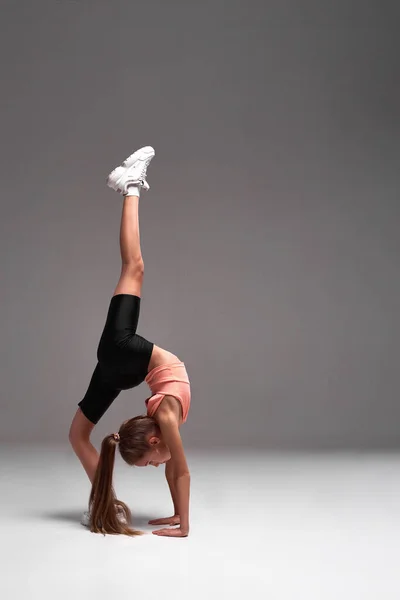 Get active. Flexible cute little girl child bending over backwards while stretching her body isolated on a grey background. Sport, training, fitness, active lifestyle concept. Vertical shot. — Stock Photo, Image