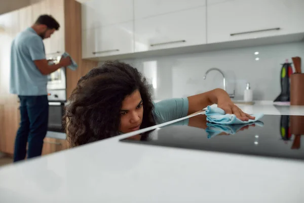 Ci occuperemo noi del casino. Giovane donna afro-americana con viso serio pulire stufa elettrica con strofinare tessile mentre si lavora con il suo collega di lavoro maschile nella cucina moderna. Concetto di servizi di pulizia — Foto Stock