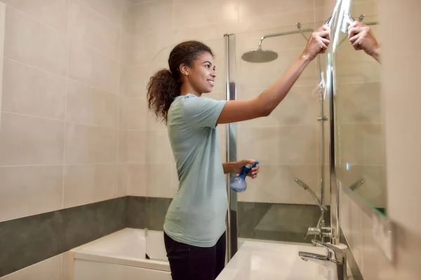 Odrobina perfekcji. Młoda uśmiechnięta afro amerykańska kobieta w mundurze czyści lustro za pomocą squeegee i detergentu natryskowego podczas pracy w łazience — Zdjęcie stockowe