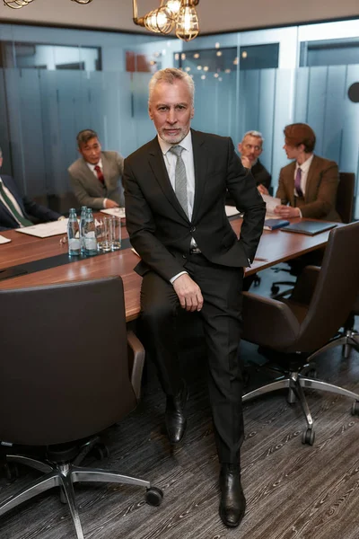 Бизнес-эксперт. Серьезный зрелый бизнесмен в формальной одежде держит одну руку в кармане и смотрит в камеру, опираясь на стол в офисе — стоковое фото