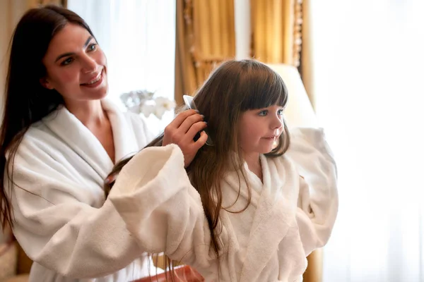 Encantadora atmósfera. Primer plano de madre e hija caucásicas relajándose en una acogedora habitación de hotel. Mamá está cepillando el pelo de los niños. El concepto de familia, amor, cuidado — Foto de Stock