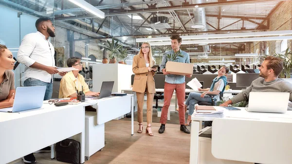 Bienvenidos a nuestro equipo. Joven hombre y mujer de pie en el centro de la oficina mientras sus nuevos compañeros de trabajo se sientan a la mesa y trabajan — Foto de Stock