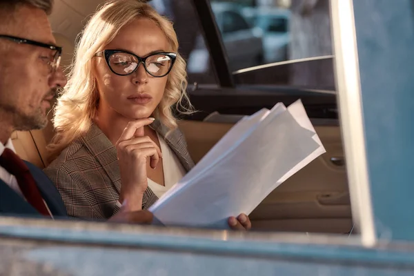 Déjeme pensar. Dos colegas en gafas con documentos y mirándolos mientras están sentados en el coche — Foto de Stock