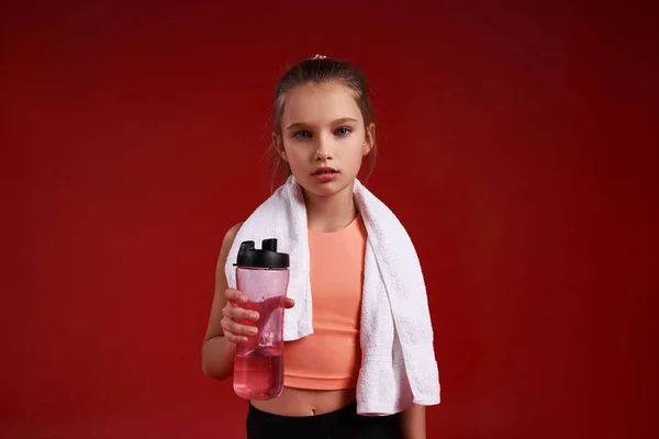 긴장풀어. 스포츠에 종사하는 귀여운 소녀는 수건 과 물병을 들고 서서 카메라를 보고 있습니다. 빨간 배경에 고립되어 있습니다. 건강, 훈련, 활동적 인 생활 방식의 개념. 수평 샷 — 스톡 사진
