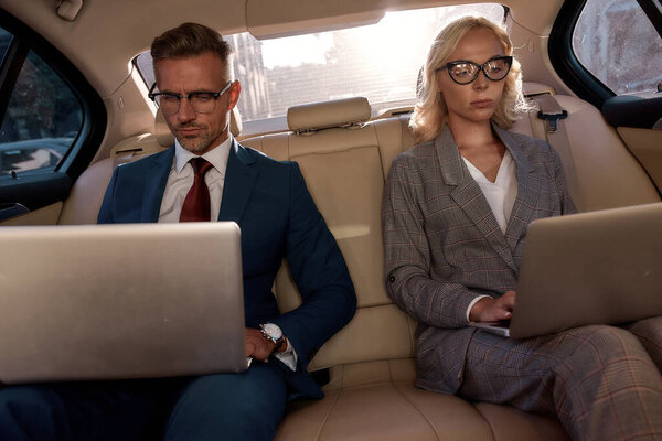 Business professionals. Two colleagues in classic wear working on their laptops while sitting in the car