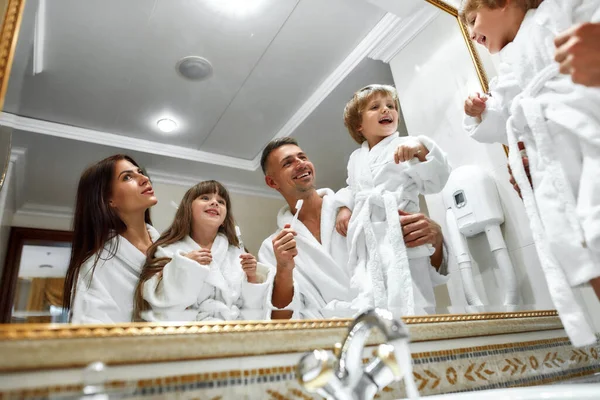 Cuidados dentários. Pais caucasianos e duas crianças em roupões de banho brancos tendo rotina matinal no banheiro do hotel, de pé perto do espelho, sorrindo depois de escovar os dentes. Conceito de férias em família — Fotografia de Stock