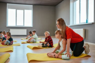 Doing butterfly exercise. Young female trainer helping little girl to stretch her legs. Group of children sitting on the floor and doing gymnastic exercises in the dance studio