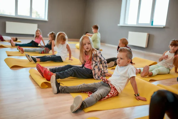 Aspetto un allenatore. Gruppo di bambini seduti su tappetini gialli da yoga nella sala da ballo. Piccoli ragazzi e ragazze che fanno esercizi ginnici — Foto Stock