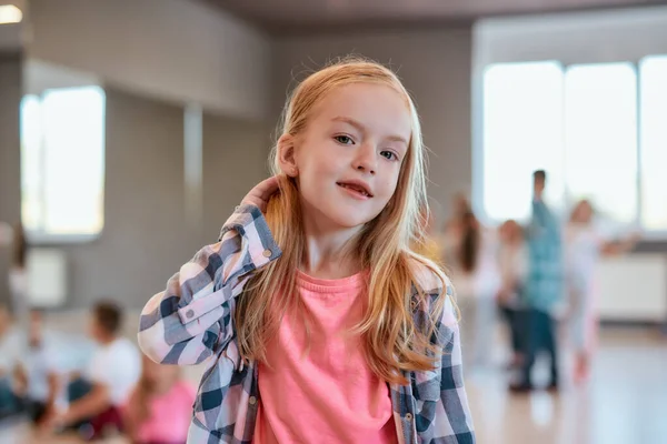 Portrait of a cute little girl in casual clothes adjusting hair and looking at camera while standing in the dance studio — 스톡 사진