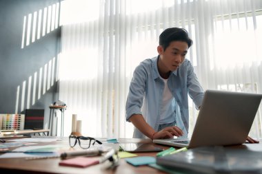 Busy designer at work. Serious asian man in casual wear looking on laptop while standing in the modern office