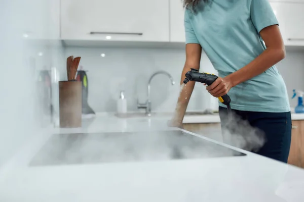 Serviço no trabalho. Vista cortada de uma mulher em uniforme de limpeza fogão elétrico com limpador de vapor enquanto trabalhava na cozinha moderna — Fotografia de Stock