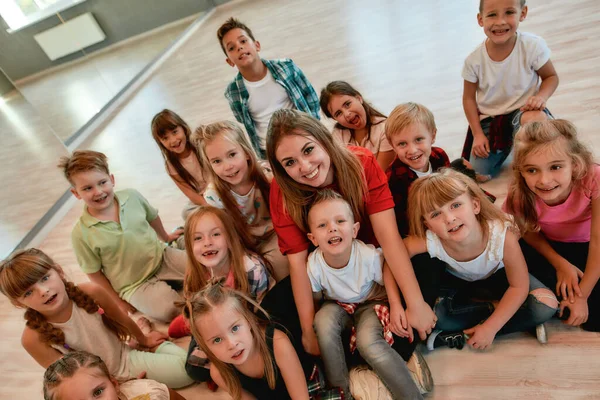Учитель и дети. Счастливая женщина-тренер по танцам с позитивными и симпатичными детьми, смотрящими в камеру и улыбающимися, сидя на полу в танцевальной студии — стоковое фото