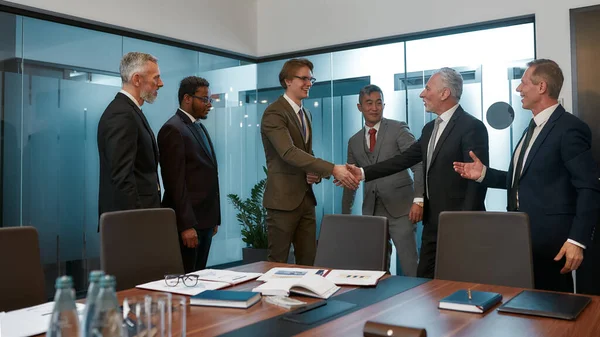 Great job. Two happy businessmen in classic wear shaking hands after signing the agreement while standing with their team in the modern office — Zdjęcie stockowe