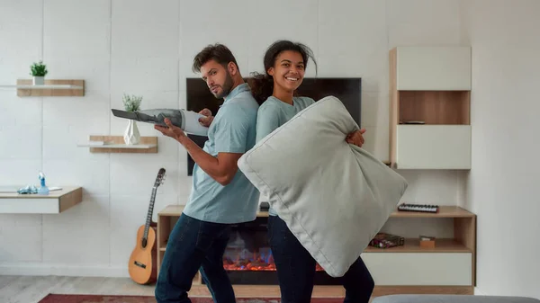 Having fun. Two young professional cleaners having fun while working together in the living room. Young caucasian man playing with handheld vacuum, afro american woman holding sofa pillow and smiling — Zdjęcie stockowe
