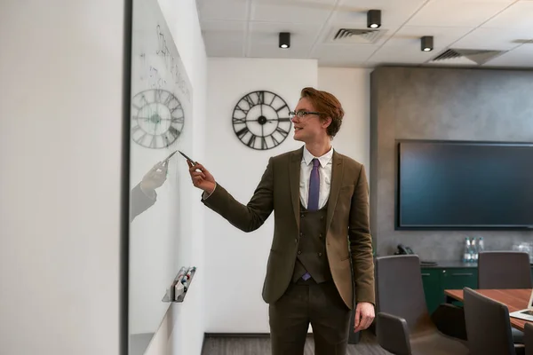 Great result. Portrait of young cheerful businessman in classic wear pointing at white blackboard and explaining something to his colleagues while having a meeting. Presentation — Φωτογραφία Αρχείου