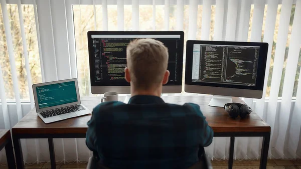 Code propre. Vue arrière du jeune développeur web masculin caucasien écrivant le code du programme tout en étant assis sur le lieu de travail avec trois moniteurs dans le bureau moderne. Programmation Web — Photo