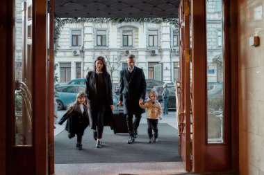 Business trip. Full-length shot of family of tourists, father, mother, daughter and son in casual wear with a grey suitcase entering the hotel.