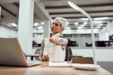 Exhaustion. Young attractive tattooed business woman feeling tired and massaging her neck while sitting at her working place in the modern office