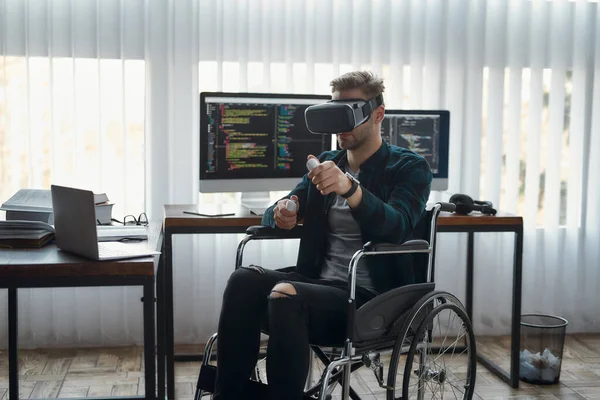 Tecnologías que cambian el mundo. Joven desarrollador web masculino en silla de ruedas con auriculares de realidad virtual mientras trabaja en la oficina moderna. Múltiples pantallas de ordenador con código de programa —  Fotos de Stock