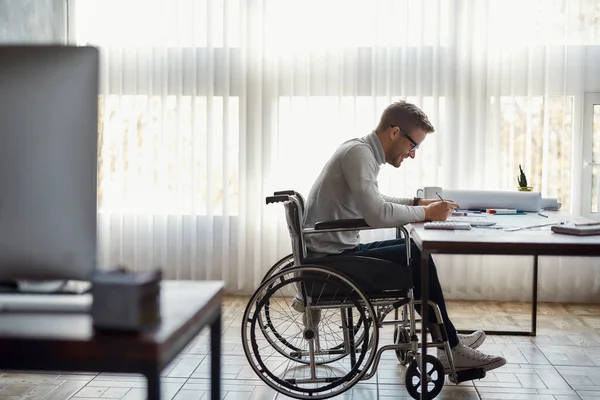 Great working day. Side view of cheerful male office worker in a wheelchair drawing something and smiling while working at his workplace in the modern office — Stockfoto