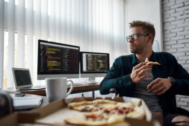 Taking a break. Portrait of hungry male web developer in a wheelchair eating pizza while sitting at his workplace in the modern office. Multiple computer screens with program code