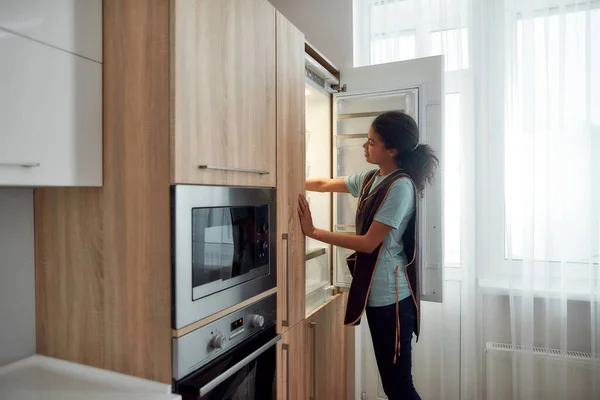 Your home is in good hands. Young afro american house maid in uniform cleaning refrigerator in the modern kitchen. Cleaning services — стокове фото