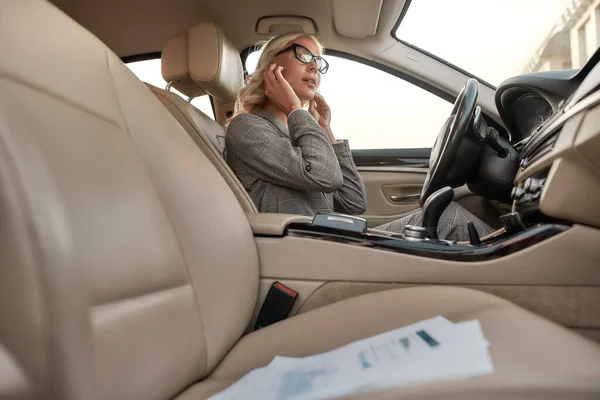 Eu devia estar sempre disponível. Empresária atraente e elegante no desgaste clássico ajustando fones de ouvido enquanto sentado em motoristas assento no carro — Fotografia de Stock