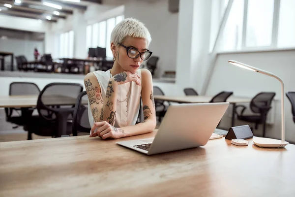 Busy day. Portrait of focused blonde tattooed businesswoman in eyeglasses looking at laptop screen while working in the modern working space — Stock Fotó