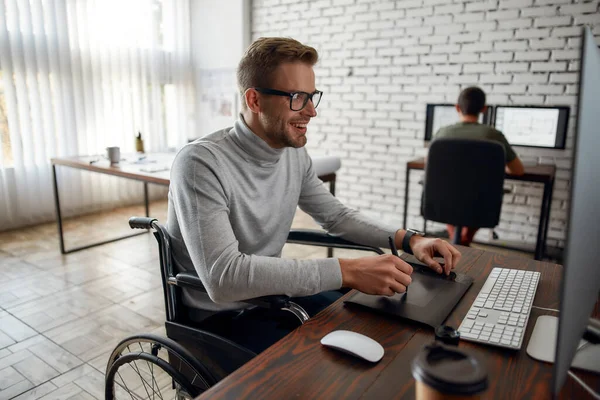 Enjoying his work. Side view of young and positive male office worker in a wheelchair using stylus pen and digital tablet while working in co-working space — Stockfoto