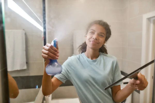 Sprzątanie miłością. Młoda, uśmiechnięta afro amerykańska kobieta w mundurowych drzwiach prysznicowych czyszczących z wycieraczką i detergentem natryskowym — Zdjęcie stockowe
