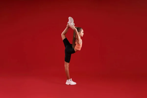 Excellence. A cute kid, girl is engaged in sport, gymnast is looking aside while stretching her legs. Isolated on red background. Fitness, training, active lifestyle concept. Horizontal shot — Stockfoto