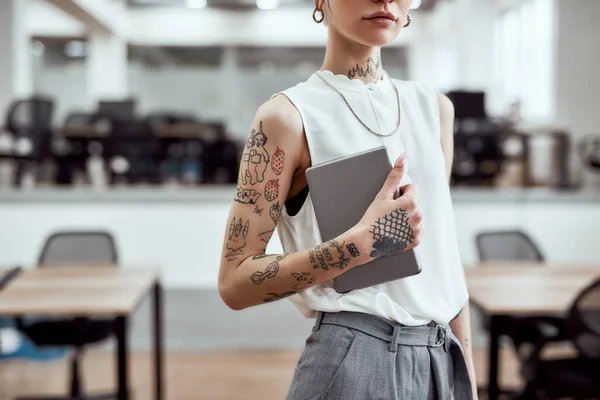 At work. Cropped photo of young stylish business woman with tattoos on her arm holding digital tablet while standing in the modern working space — 스톡 사진
