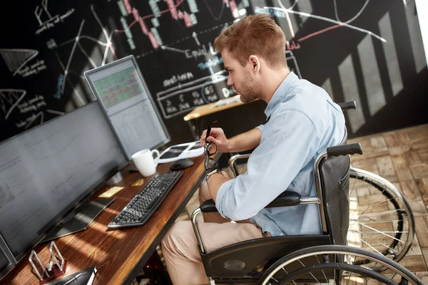 Successful investor. Side view of young male trader in a wheelchair working with charts and market reports on multiple computer screens while sitting against blackboard in the modern office — Stok fotoğraf