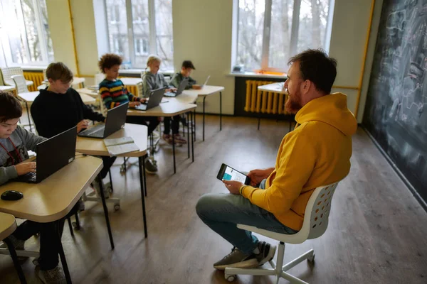 Where Learning Begins. Portrait of young male teacher communicating with his pupils, while sitting with tablet pc near the blackboard during a lesson in modern smart school — Φωτογραφία Αρχείου