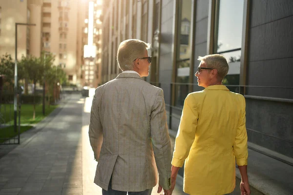 Walking with You. Back view of modern middle-aged couple holding hands and looking at each other while walking together through the city street — Stockfoto