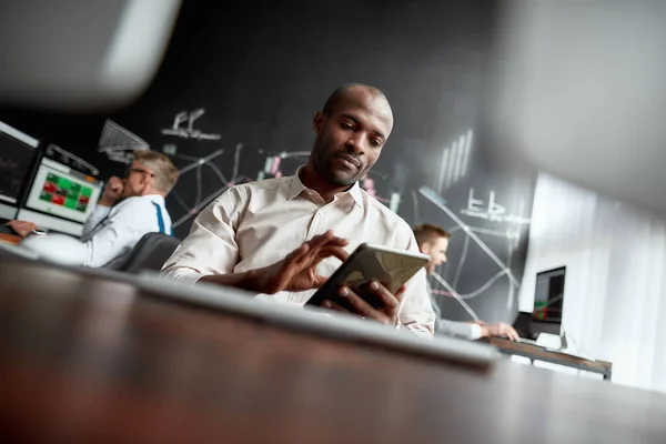 De beste financiële gemoedstoestand. Gericht Afrikaans mannelijk handelaar zitten aan het bureau en het bestuderen van analytische rapporten met behulp van tablet pc in het kantoor. Zijn collega 's werken op de achtergrond — Stockfoto