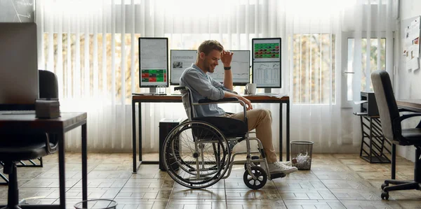Great results. Side view of cheerful young male trader worker in a wheelchair feeling happy while working at workplace with three computer monitors in the office. Disability and handicap concept — Stock Photo, Image