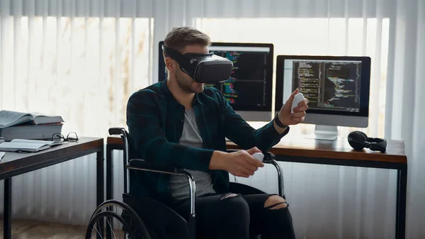 Una herramienta poderosa. Retrato de un joven desarrollador web masculino en una silla de ruedas con auriculares de realidad virtual mientras trabajaba en la oficina moderna. Múltiples pantallas de ordenador con código de programa. Alta tecnología —  Fotos de Stock