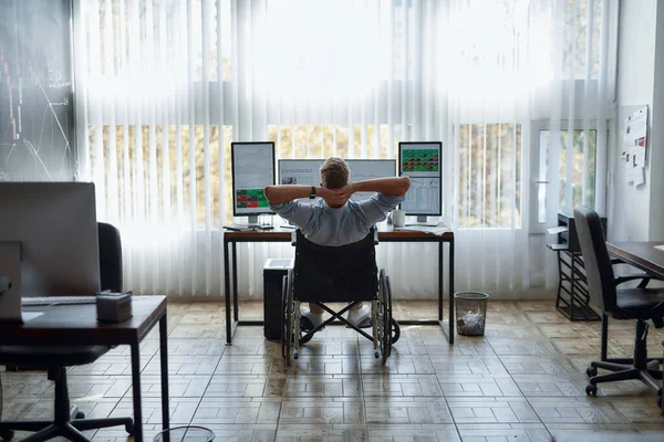 Vista posterior de un joven comerciante relajado en una silla de ruedas manteniendo las manos detrás de la cabeza y mirando a tres monitores de computadora con diagramas y gráficos mientras trabaja en la oficina moderna —  Fotos de Stock