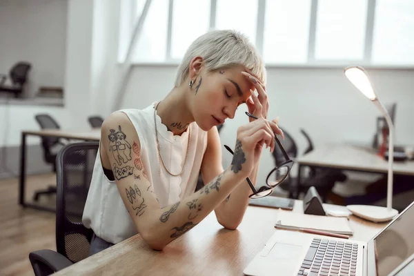 Stressful day. Portrait of tired young tattooed business woman keeping eyes closed and holding hand on head while sitting at her working place in the modern office