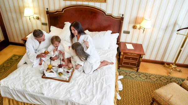 Afasta-te de tudo. Pais e duas crianças de roupão branco sorrindo enquanto tomavam café da manhã na cama, comendo e bebendo em um luxuoso quarto de hotel. Família, resort, conceito de serviço de quarto — Fotografia de Stock